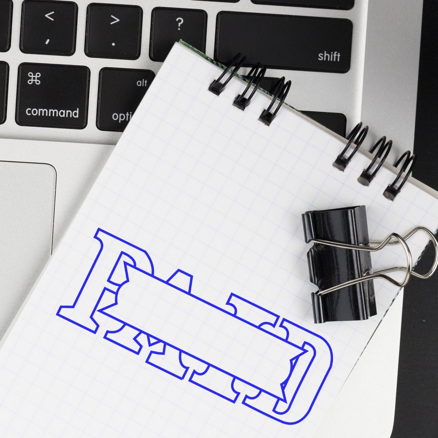 Self Inking Paid with Box Stamp used on a notepad placed on a laptop keyboard, with a black binder clip attached to the notepad.