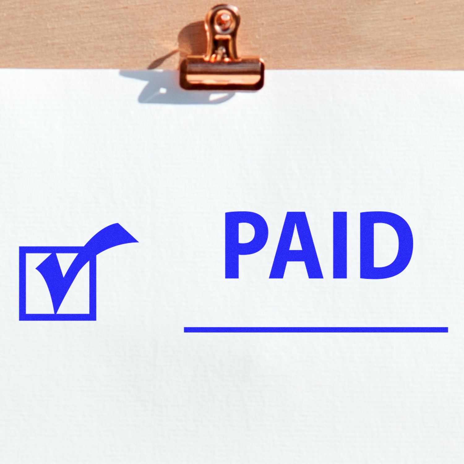 Paid with Checkmark Rubber Stamp on white paper clipped to a wooden board, featuring a blue checkmark and the word 'PAID'.