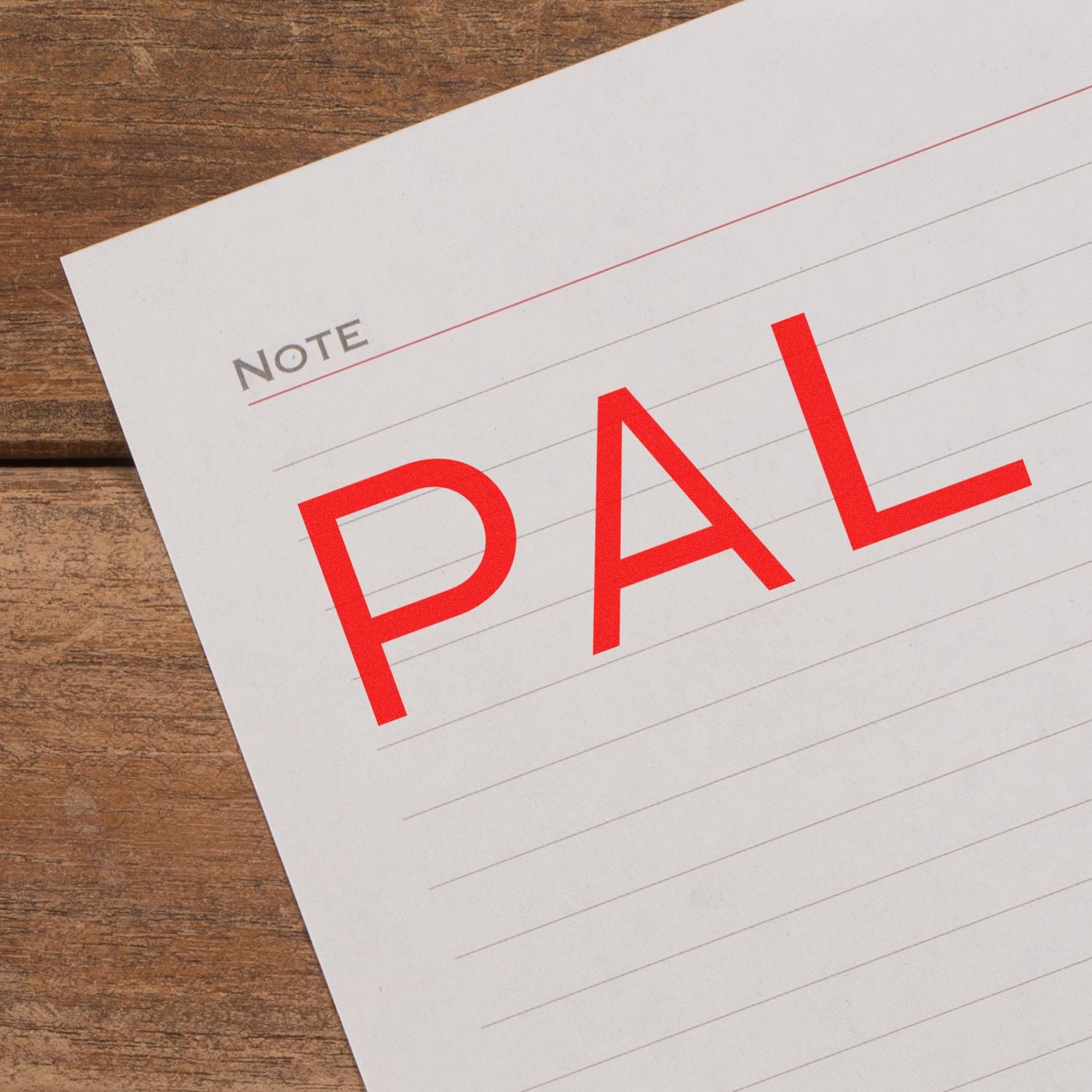 A piece of lined paper with the word PAL stamped in large red letters using the Large PAL Rubber Stamp, placed on a wooden surface.