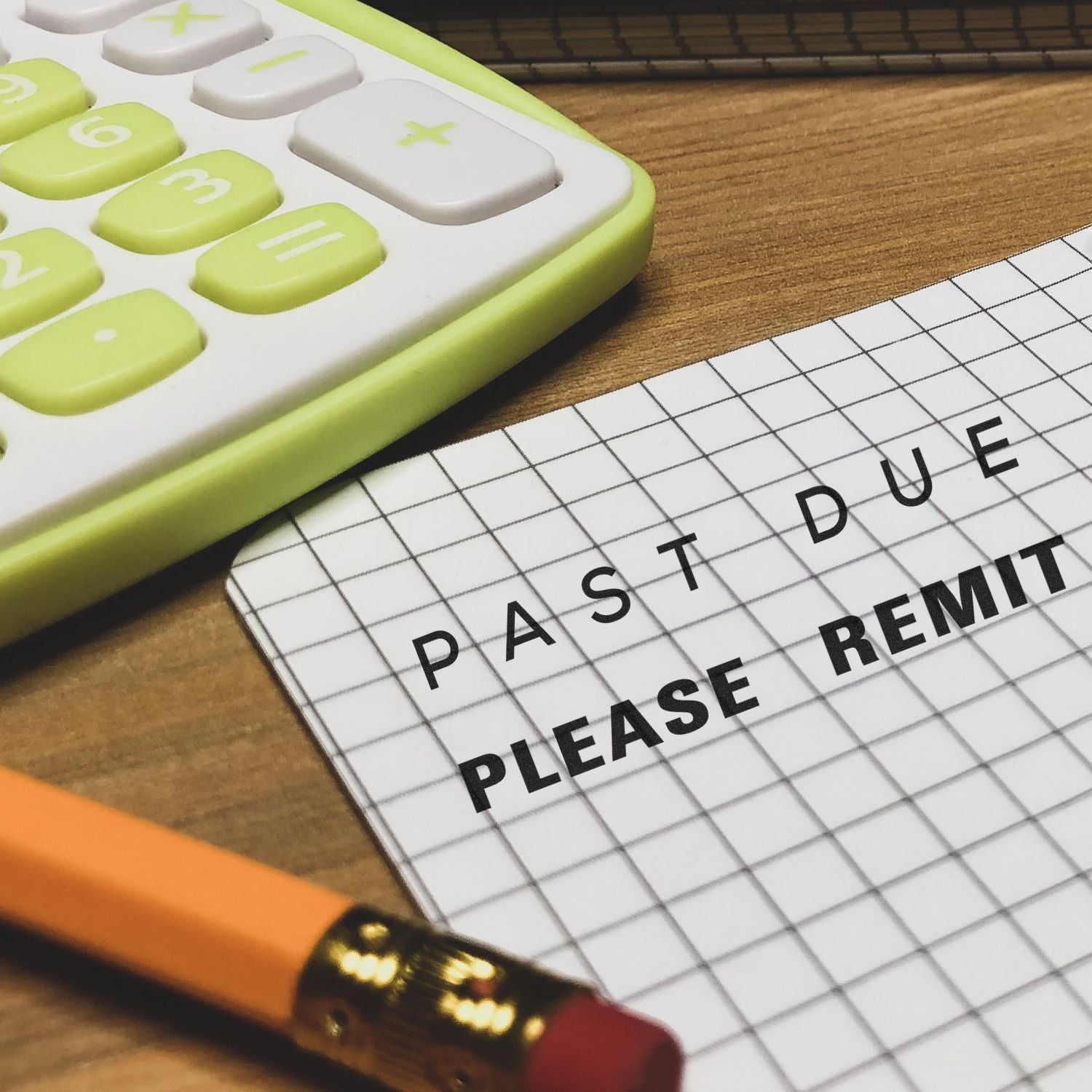 A Large Past Due Please Remit Rubber Stamp is used on a grid paper next to a yellow calculator and a pencil on a wooden desk.