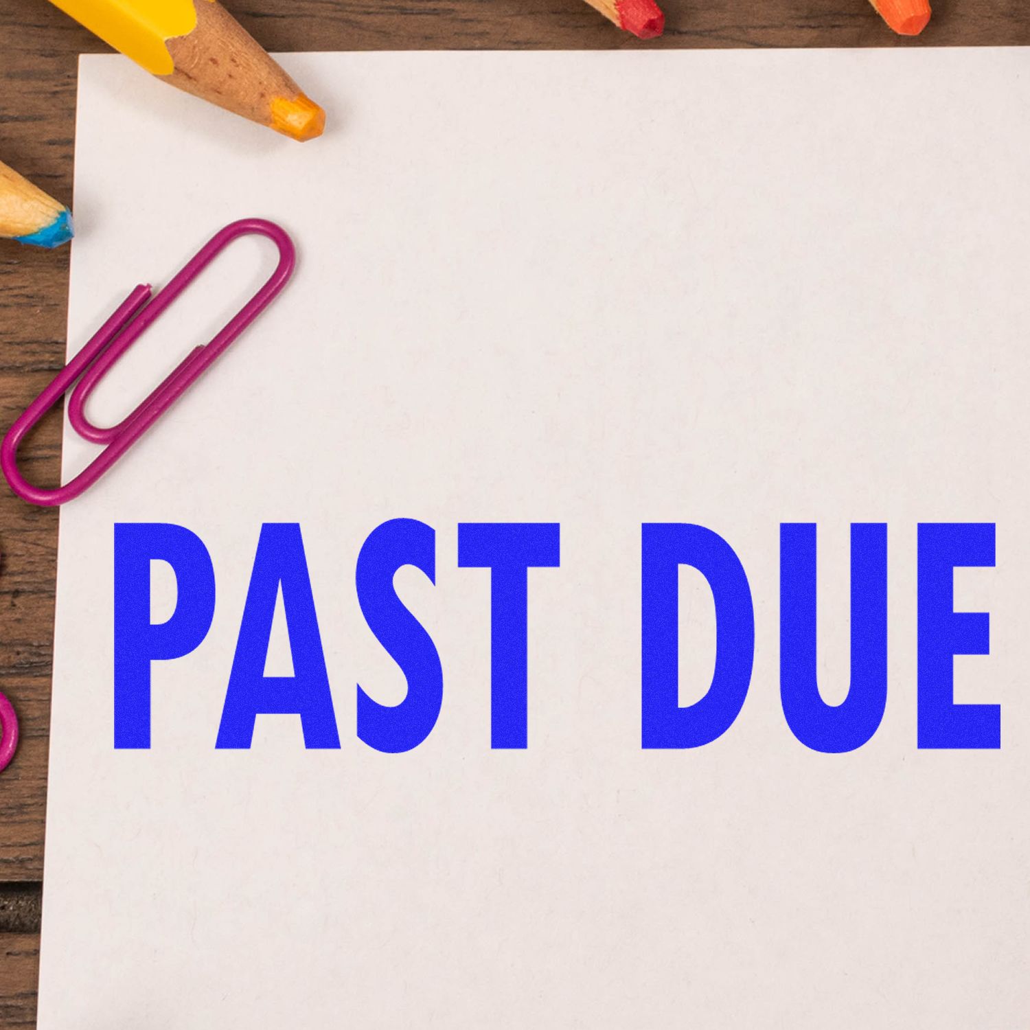 Past Due Rubber Stamp in blue ink on white paper, surrounded by colorful pencils and a pink paperclip on a wooden desk.