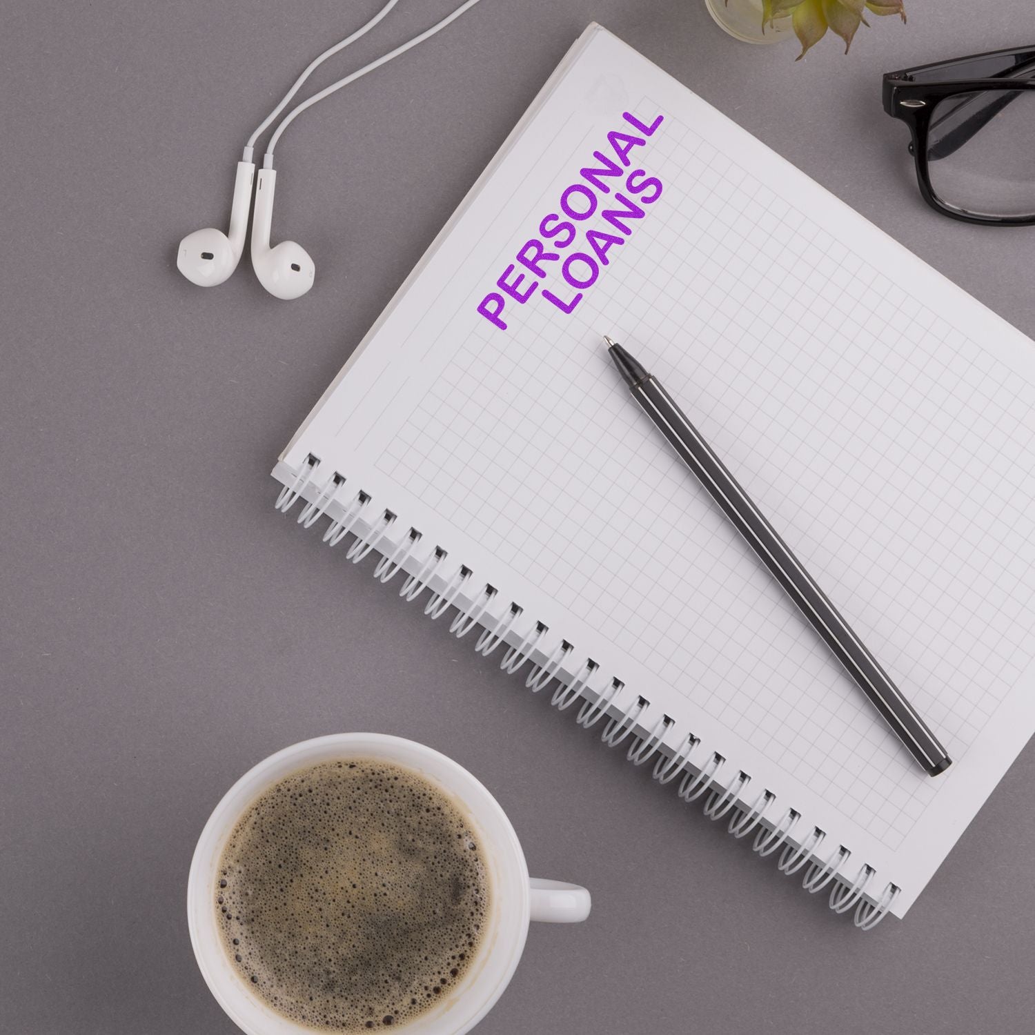 Notebook with 'Personal Loans' stamped on it, pen, coffee cup, earphones, and glasses on a gray desk.