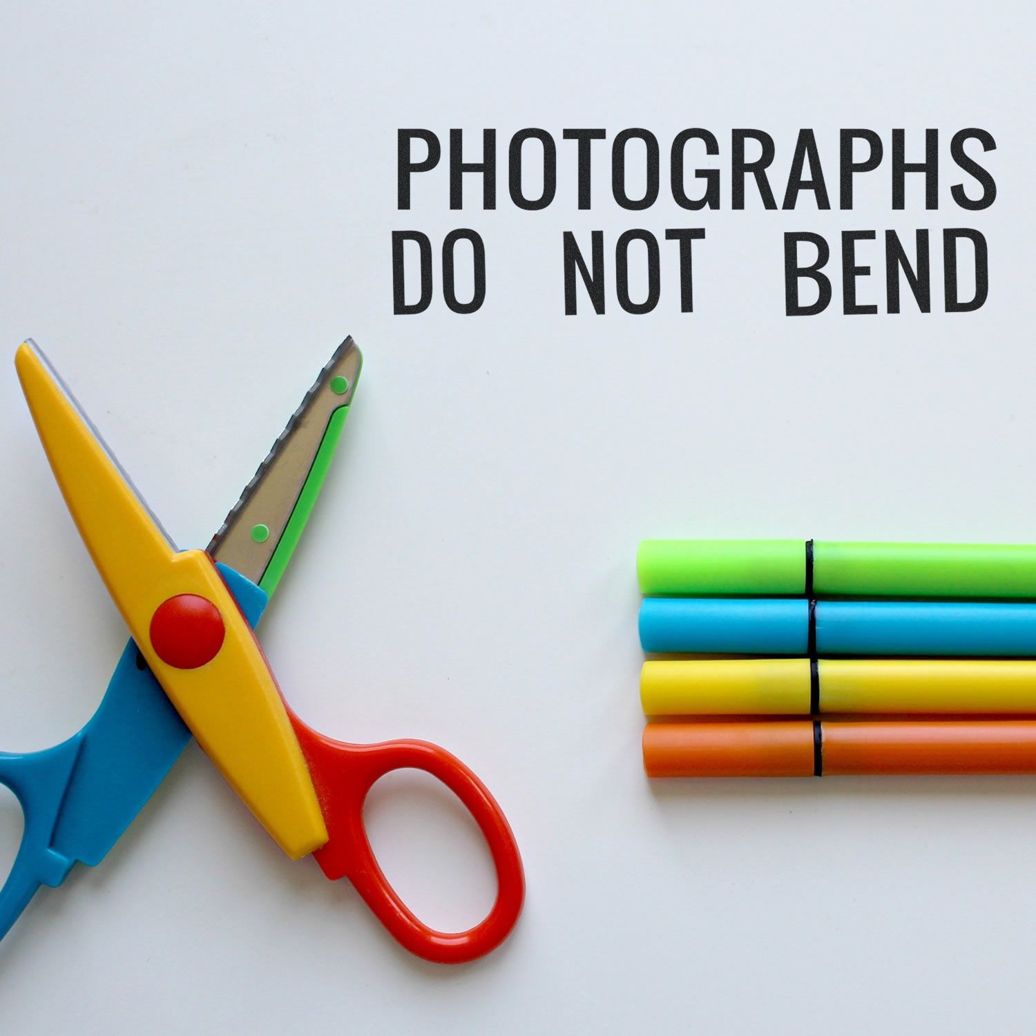 Photographs Do Not Bend rubber stamp impression on white paper, next to colorful scissors and four markers.