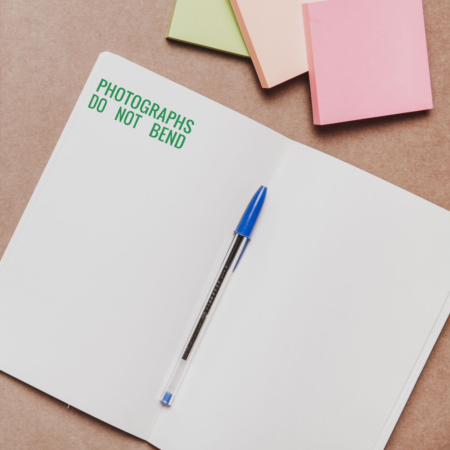 Open notebook with Photographs Do Not Bend rubber stamp in green ink, blue pen, and colorful sticky notes on a brown surface.