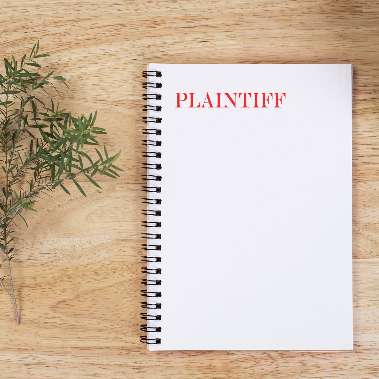 Slim Pre-Inked Plaintiff Stamp in red ink on a white spiral notebook, placed on a wooden surface next to a green plant.