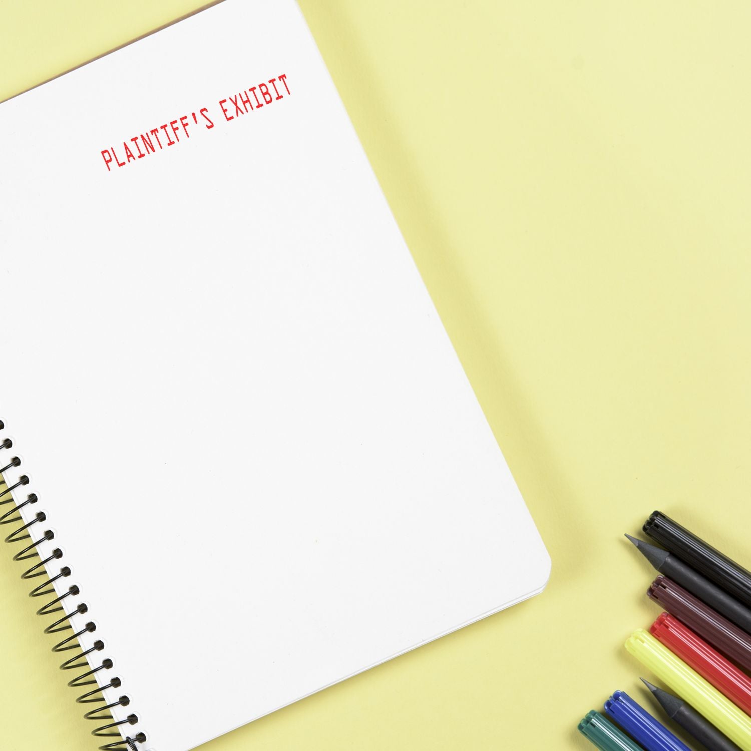 Large Self Inking Plaintiffs Exhibit Stamp marking PLAINTIFF'S EXHIBIT in red on a white notebook, with colored pens nearby on a yellow surface.