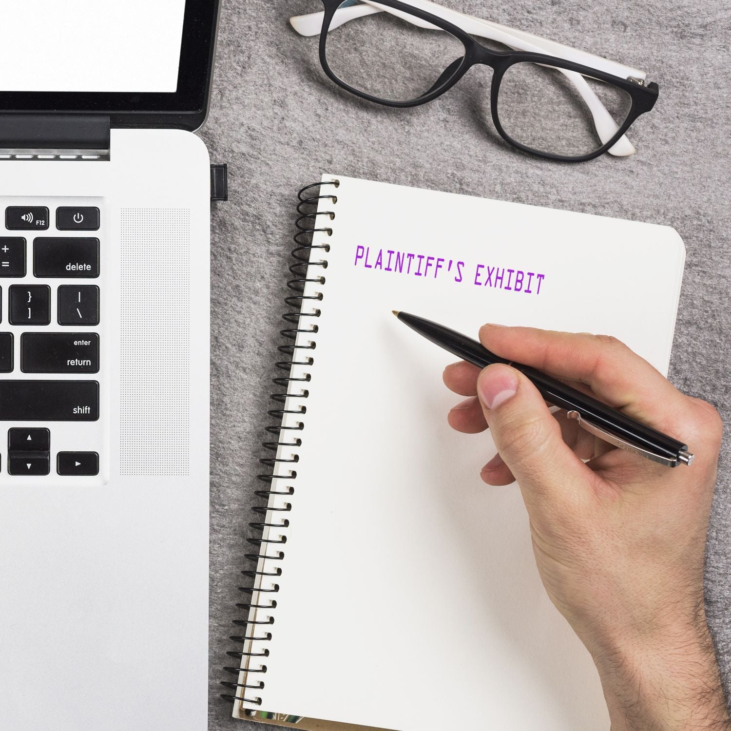 A hand holding a pen near a notebook stamped with Plaintiff's Exhibit beside a laptop and glasses on a gray surface.