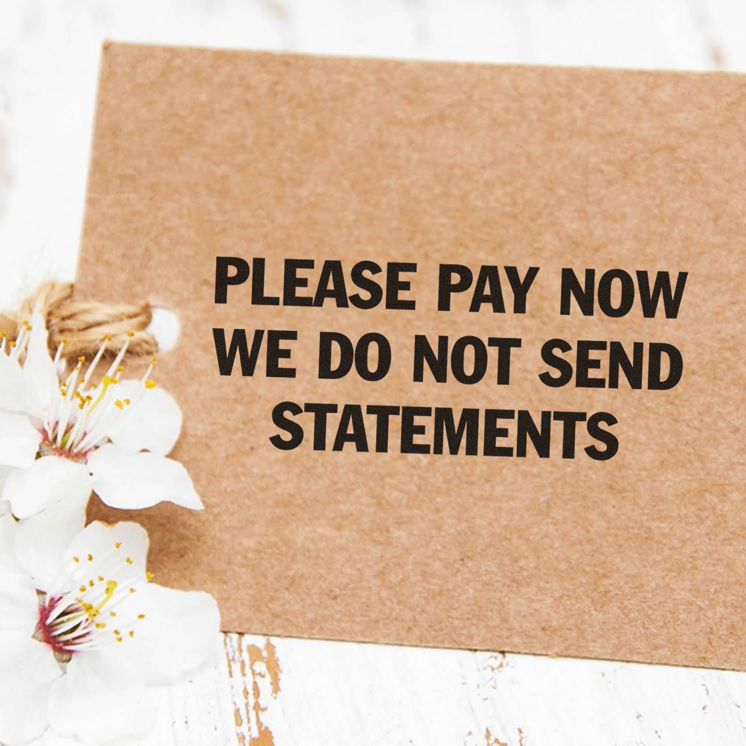 Please Pay Now No Statements Rubber Stamp on a brown card with white flowers on a rustic white wooden surface.