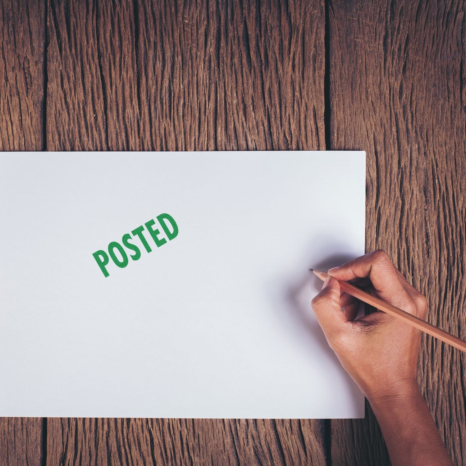 A hand holding a pencil next to a white paper stamped with POSTED in green using the Large Self Inking Posted Stamp on a wooden surface.