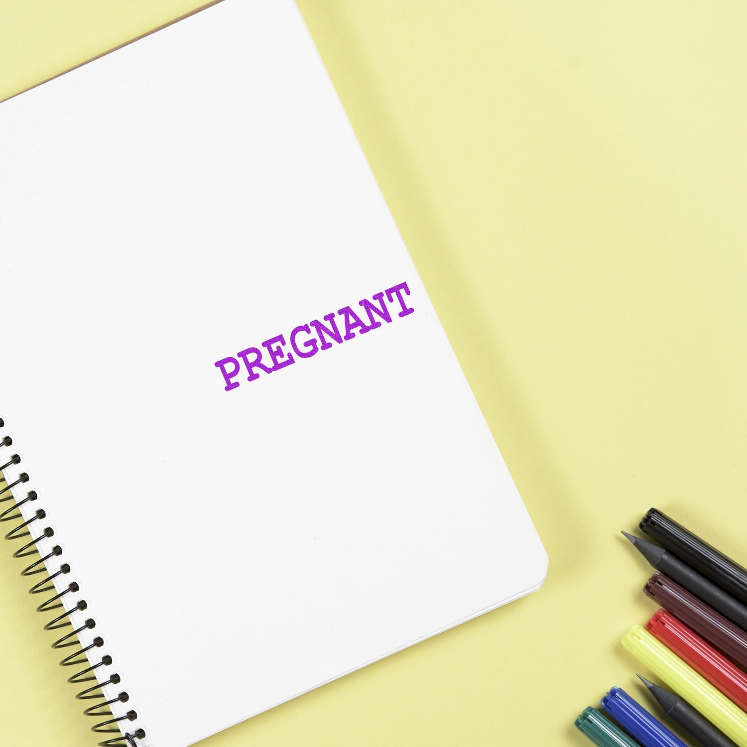 Notebook with 'Pregnant' rubber stamp in purple ink on a yellow background, surrounded by colorful pens.