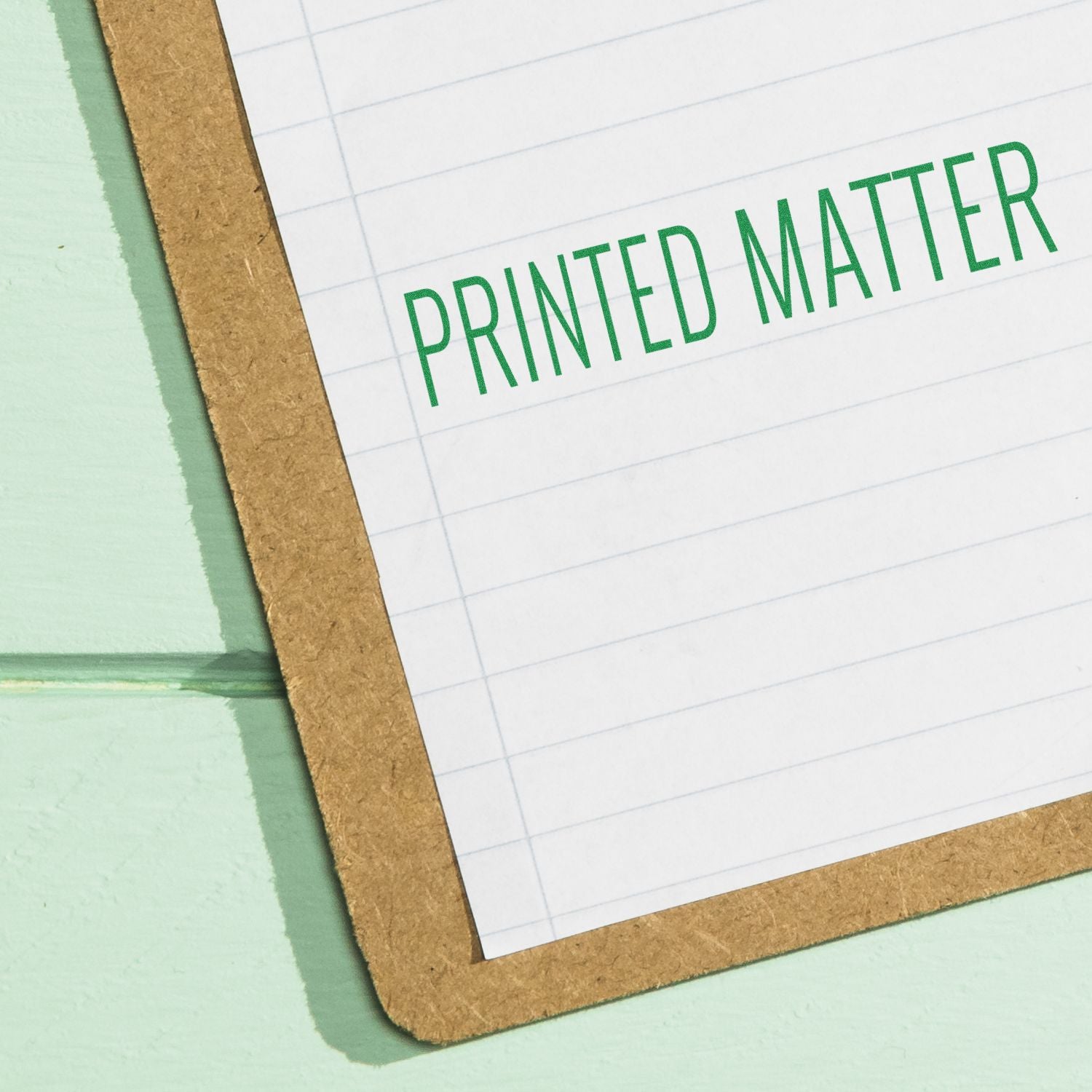 Large Self Inking Printed Matter Stamp in use, displaying PRINTED MATTER in green ink on a lined paper clipped to a brown clipboard.