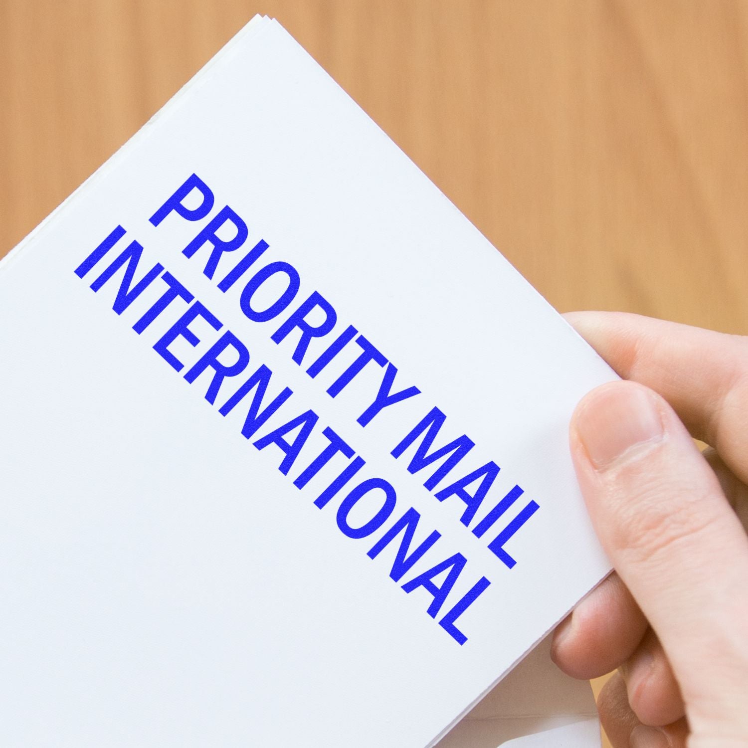 Hand holding an envelope with a blue Priority Mail International rubber stamp on a wooden background.