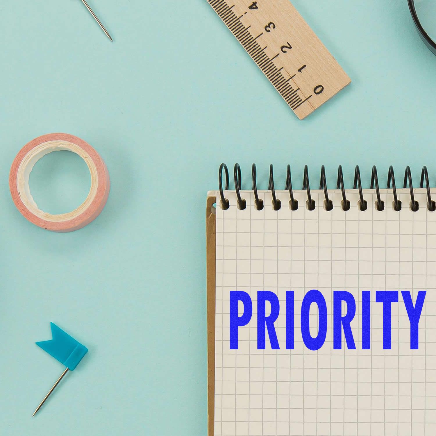 A Self Inking Priority Mailing Stamp is shown in use on a spiral notebook with the word PRIORITY stamped in blue ink, surrounded by office supplies.