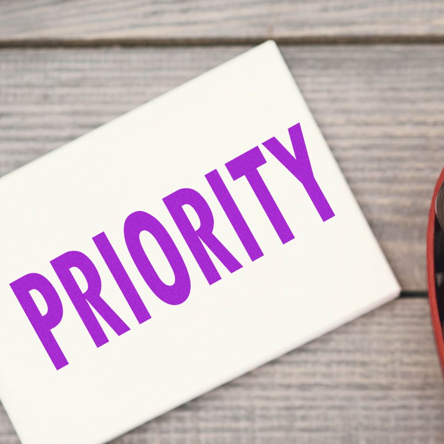 Self Inking Priority Mailing Stamp marking a white package with the word PRIORITY in bold purple letters on a wooden surface.