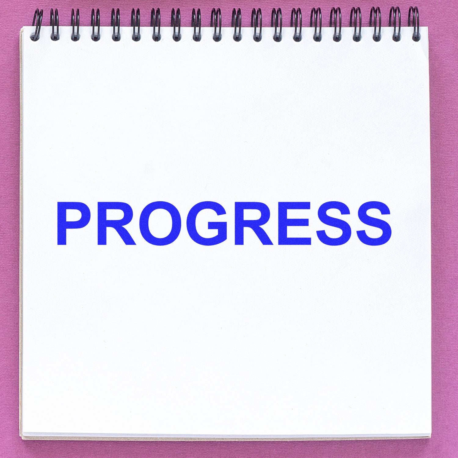 A spiral notebook with the word PROGRESS stamped in blue ink using a Self Inking Progress Stamp, placed on a pink surface.