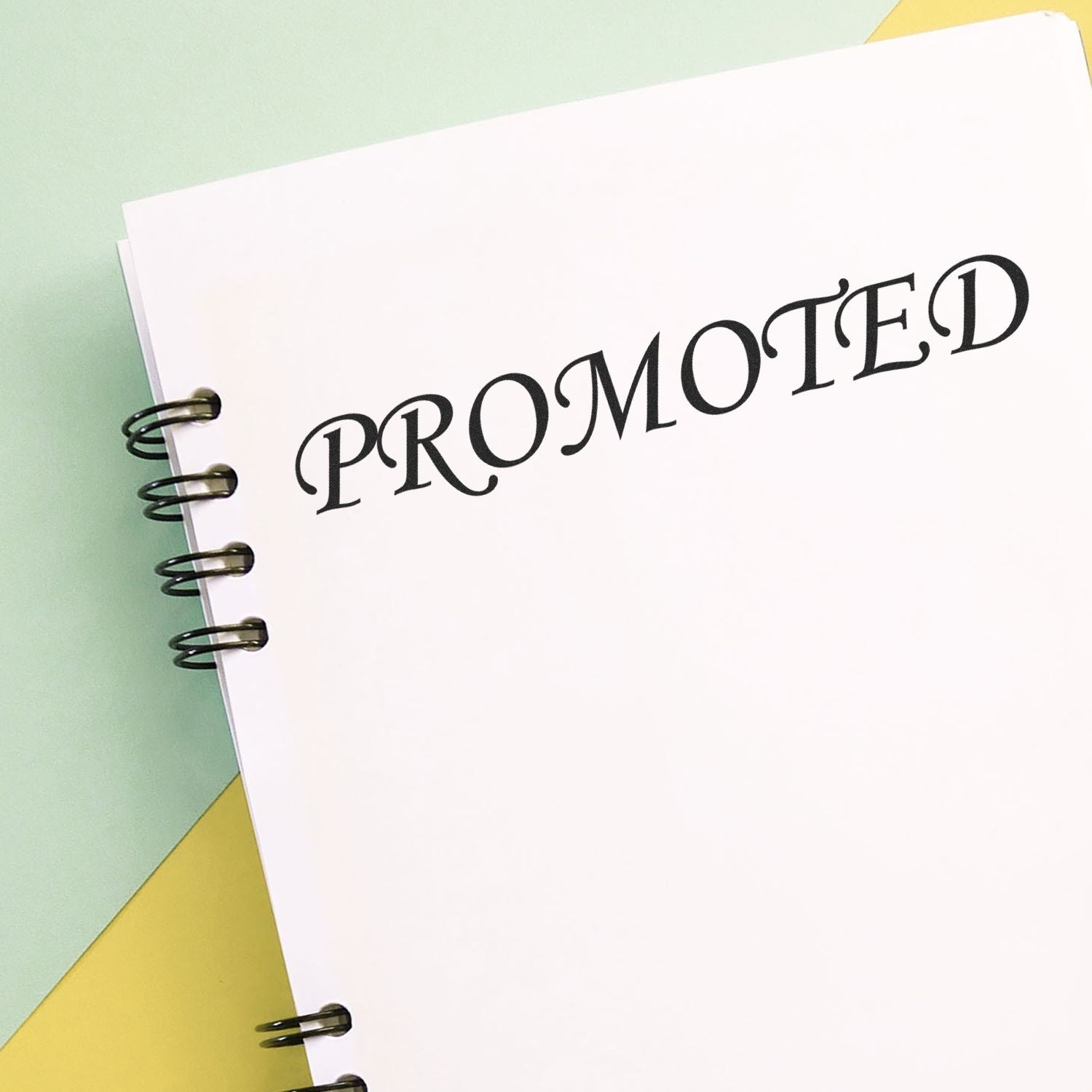 Slim Pre-Inked Promoted Stamp marking PROMOTED in bold black letters on a white spiral-bound notebook against a pastel background.