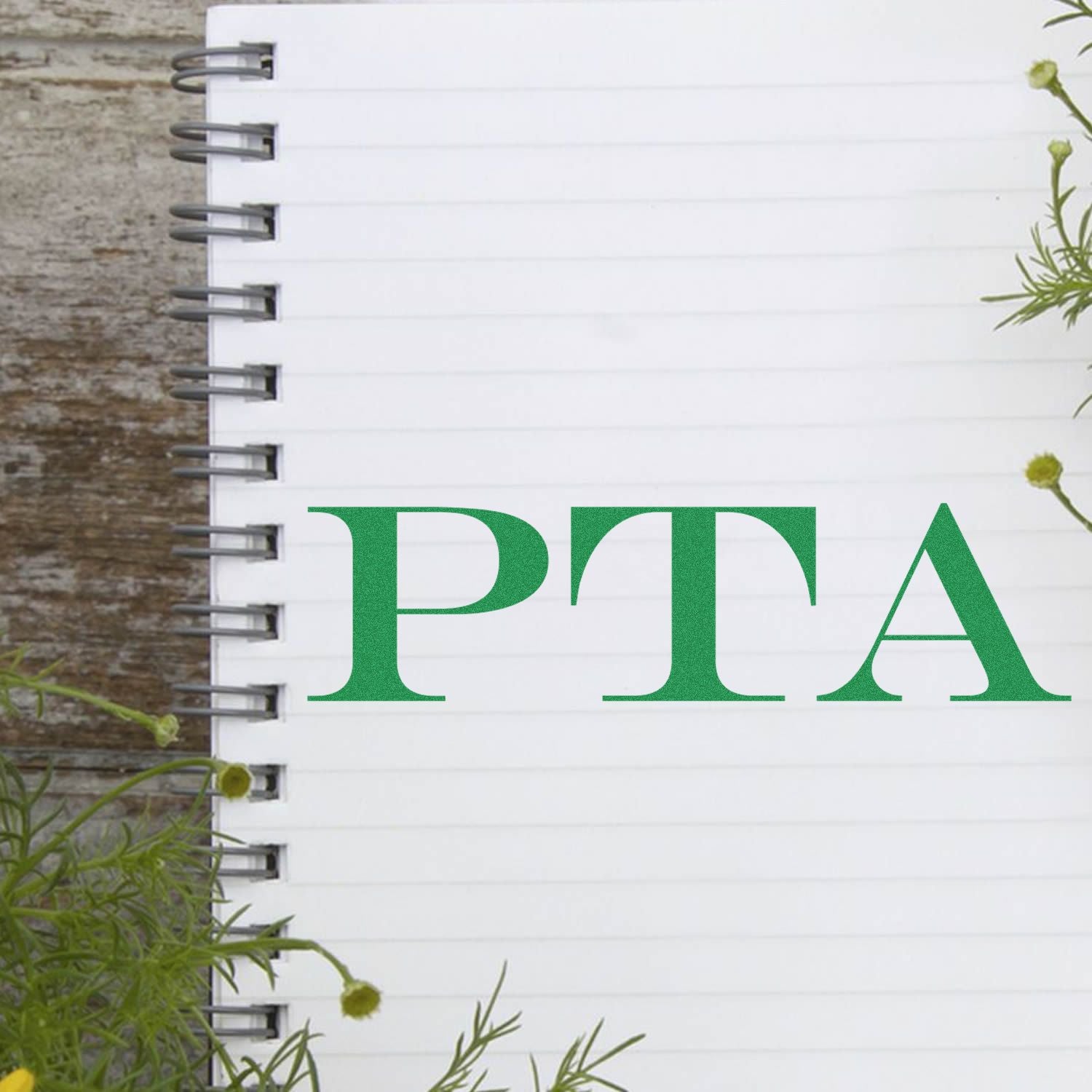 Spiral notebook with 'PTA' stamped in green using a PTA Rubber Stamp, surrounded by green plants on a wooden surface.