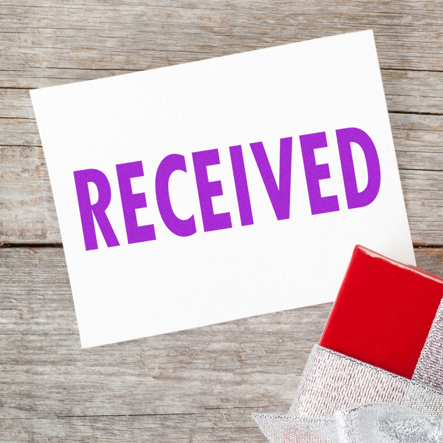 A white card with a purple RECEIVED rubber stamp on a wooden surface, next to a red and silver gift box.