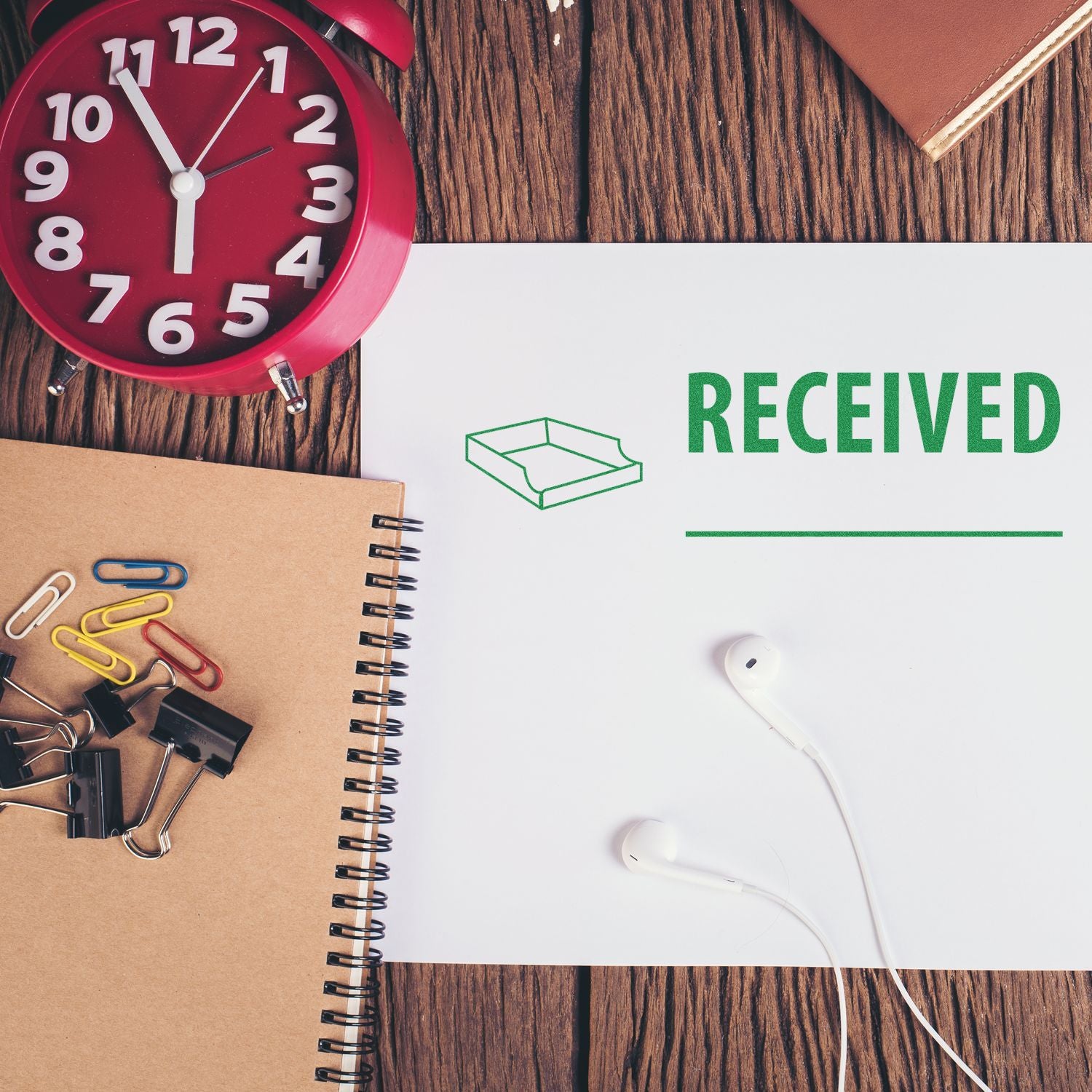 Desk with a red clock, notebook, paper clips, and a paper stamped with 'Received with Box Rubber Stamp' in green.