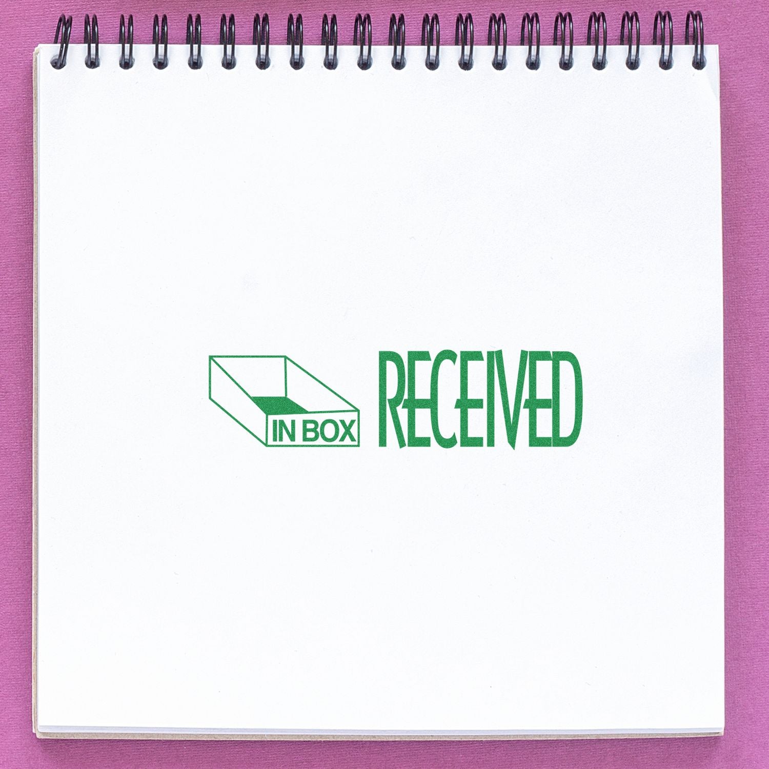 Large Self Inking Received with In Box Icon Stamp in green ink on a white spiral notebook against a pink background.