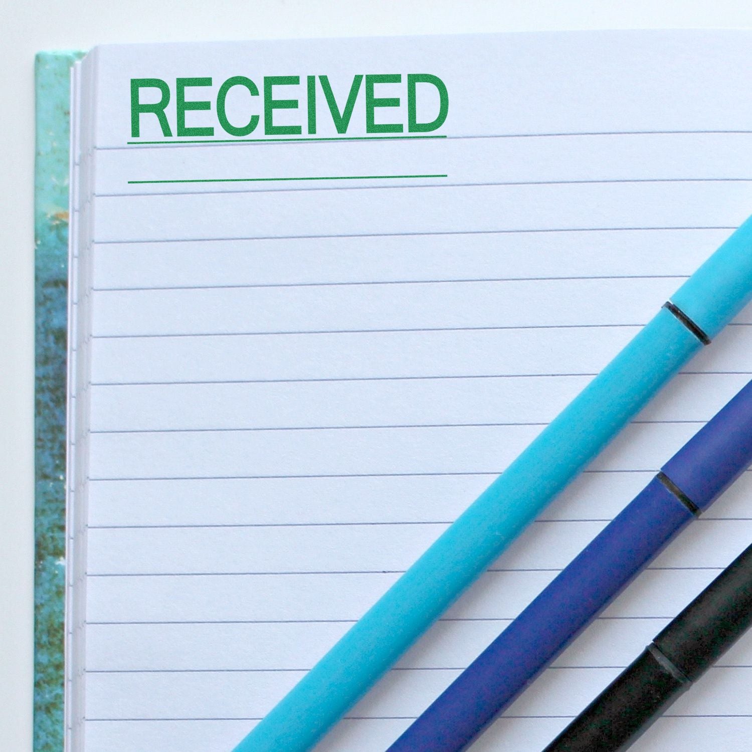 Large Self Inking Received with Line Stamp in green ink on a lined notebook, with three pens placed diagonally across the page.