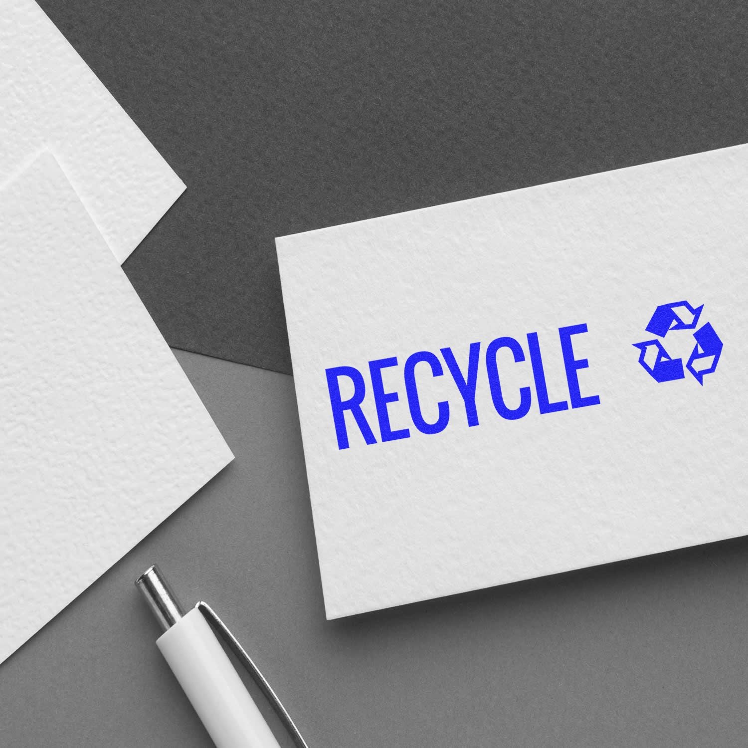 Slim Pre-Inked Recycle with Logo Stamp on white card, blue text and recycling symbol, pen and papers on gray background.