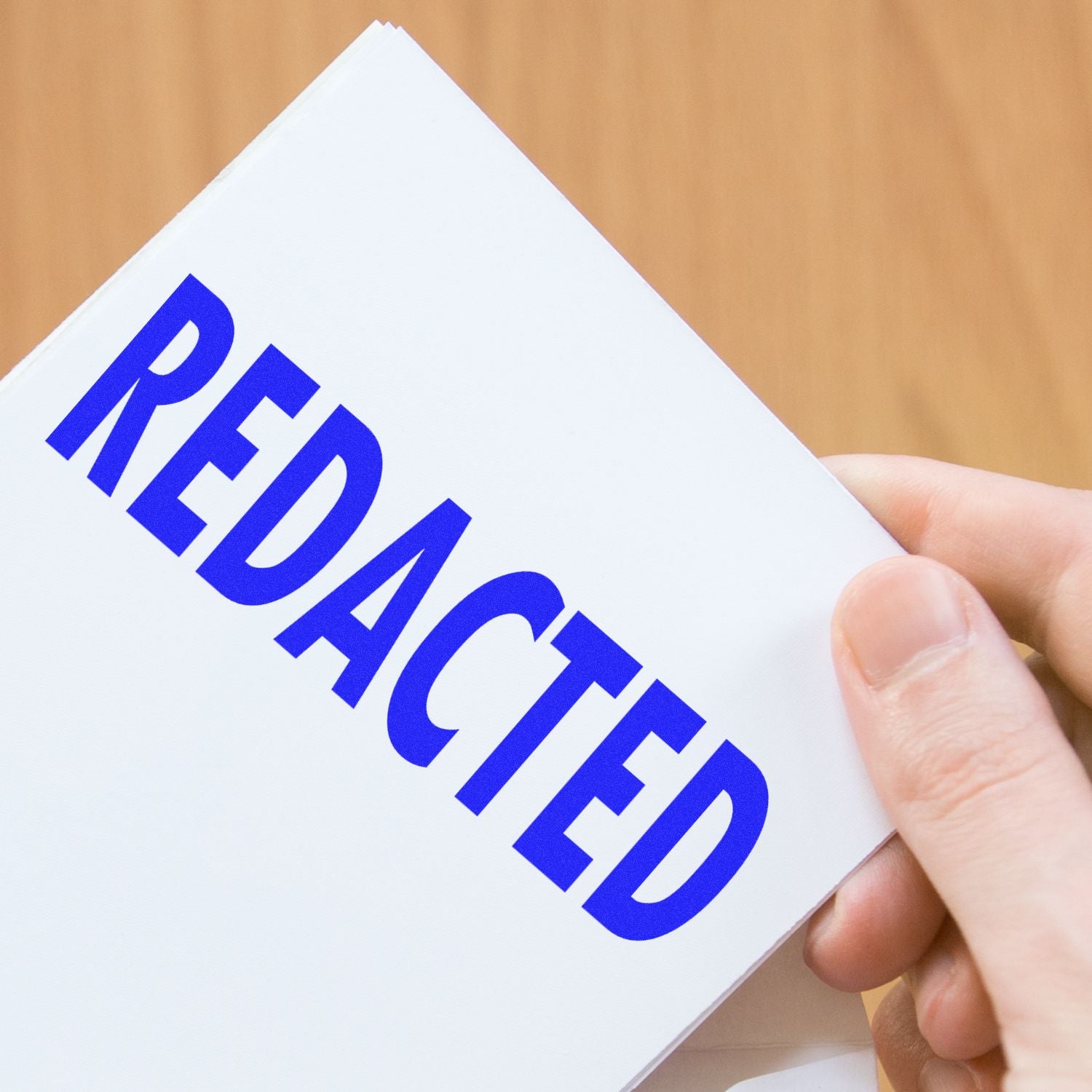 Hand holding a white paper stamped with a blue REDACTED rubber stamp, against a wooden background.