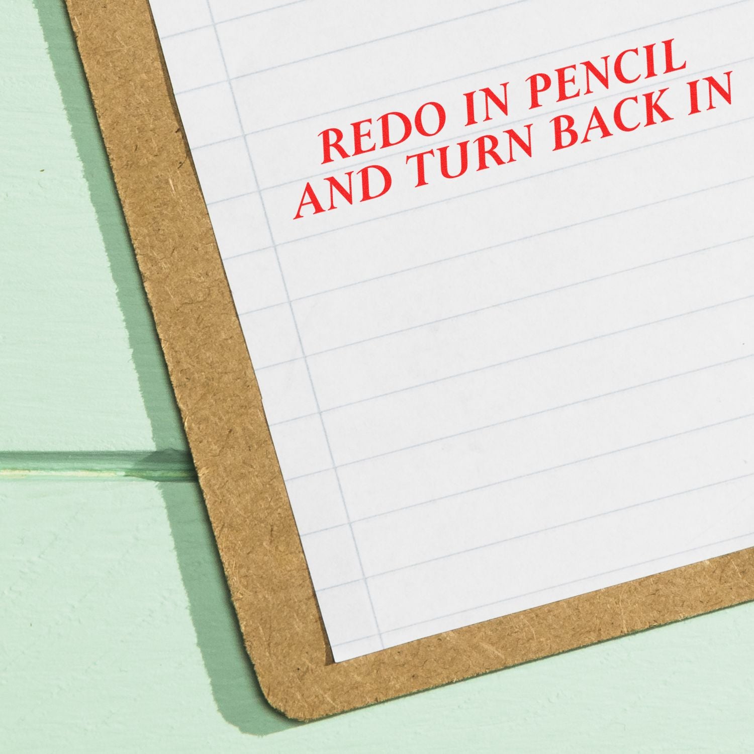 A clipboard with lined paper stamped in red ink with the words Redo In Pencil And Turn Back In Rubber Stamp on a green background.