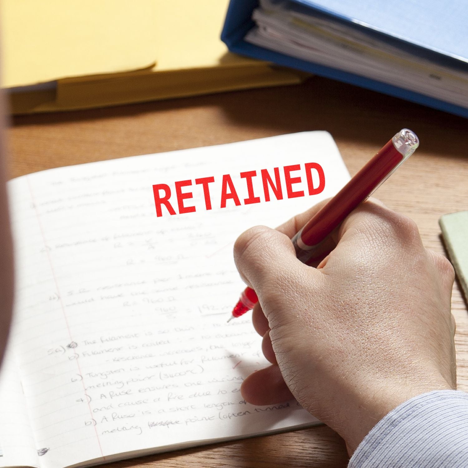 Hand using Slim Pre-Inked Retained Stamp on an open notebook with handwritten notes, highlighting the word RETAINED in red.