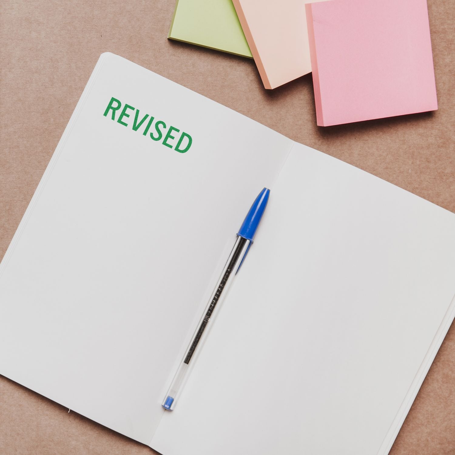 Open notebook with a blue pen and a Revised rubber stamp mark in green ink on a brown desk with colorful sticky notes.