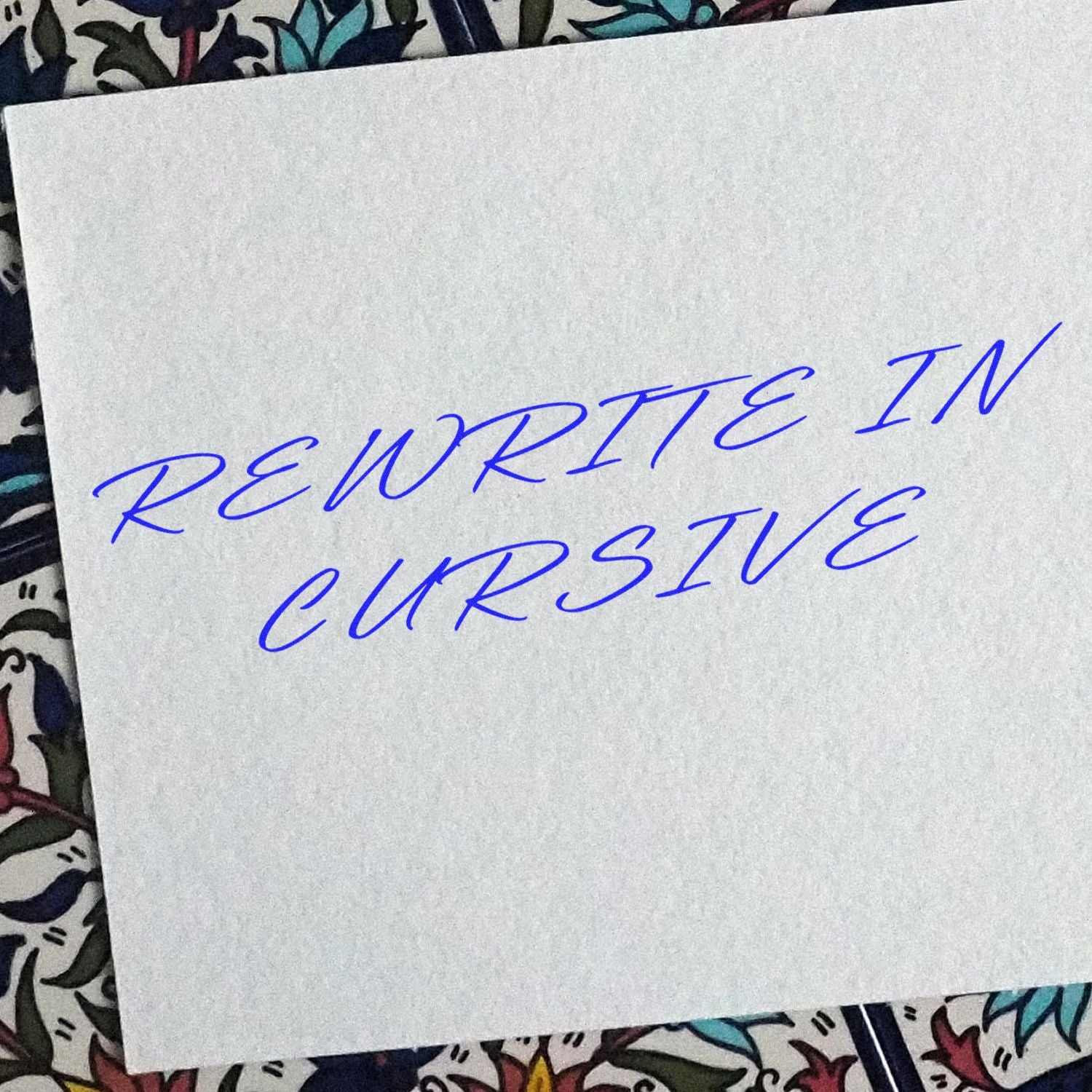 A white paper with 'Rewrite In Cursive' stamped in blue cursive letters, placed on a floral patterned background.