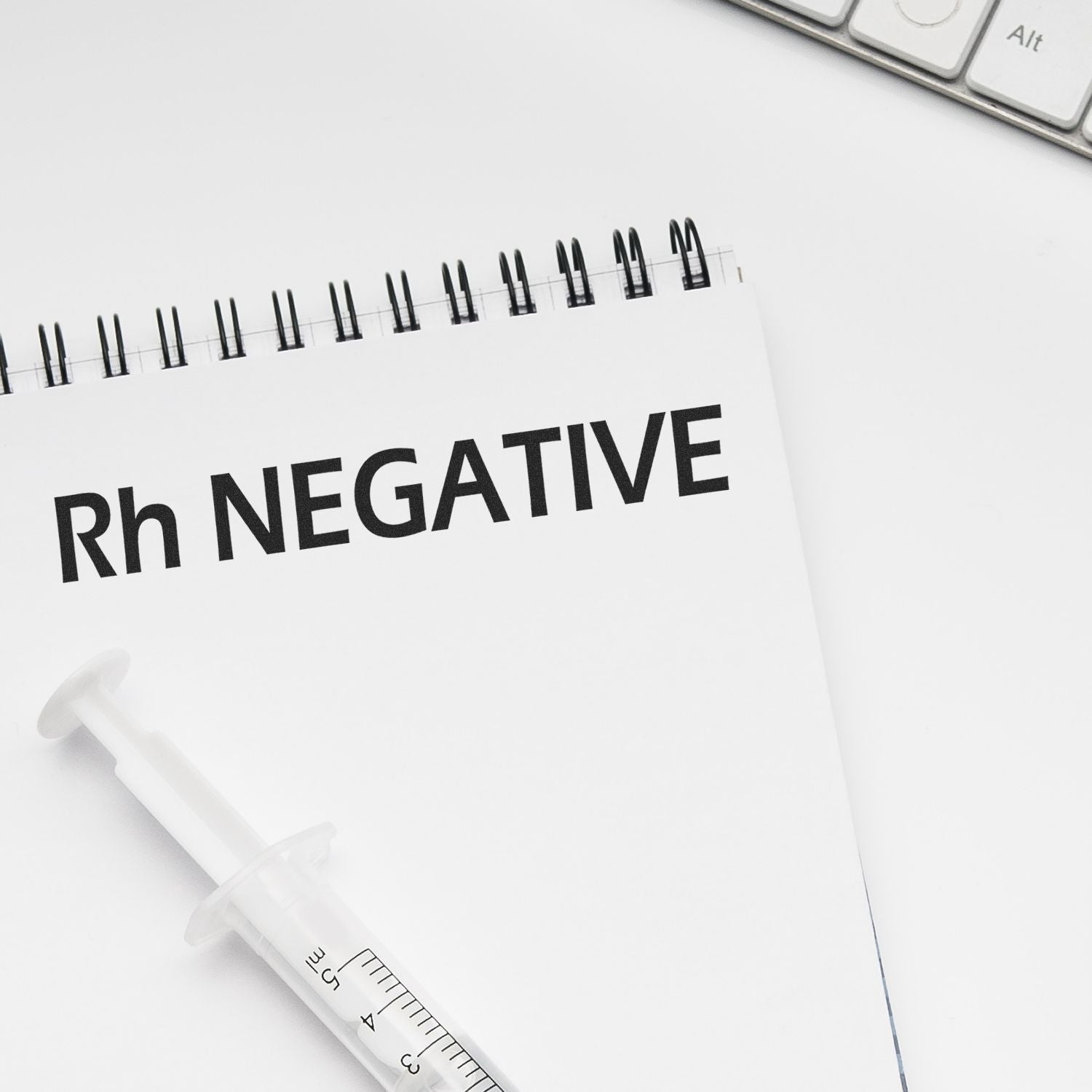 Notebook with 'Rh NEGATIVE' text, syringe, and Slim Pre-Inked Rh Negative Stamp on a white desk near a keyboard.