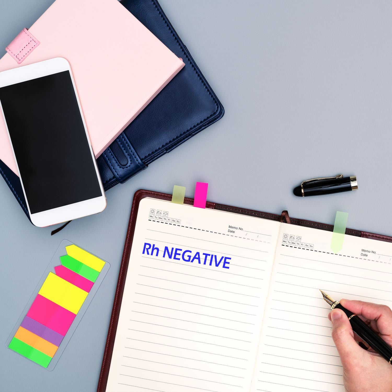 Open notebook with Rh Negative rubber stamp, colorful sticky notes, a smartphone, and a pen on a desk.