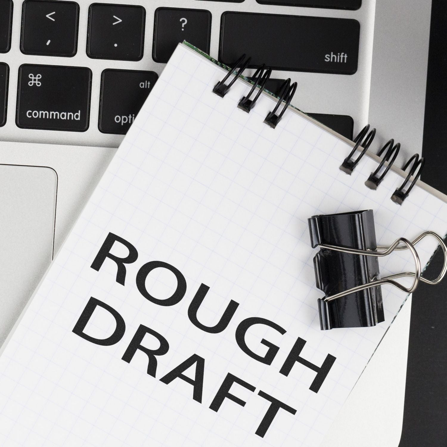 A notepad with Rough Draft stamped on it, placed on a laptop keyboard with a black binder clip attached.