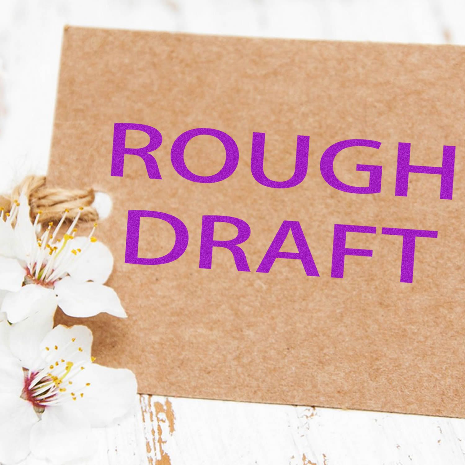 Self Inking Rough Draft Stamp used on a brown card with purple ink, placed next to white flowers on a rustic white wooden surface.