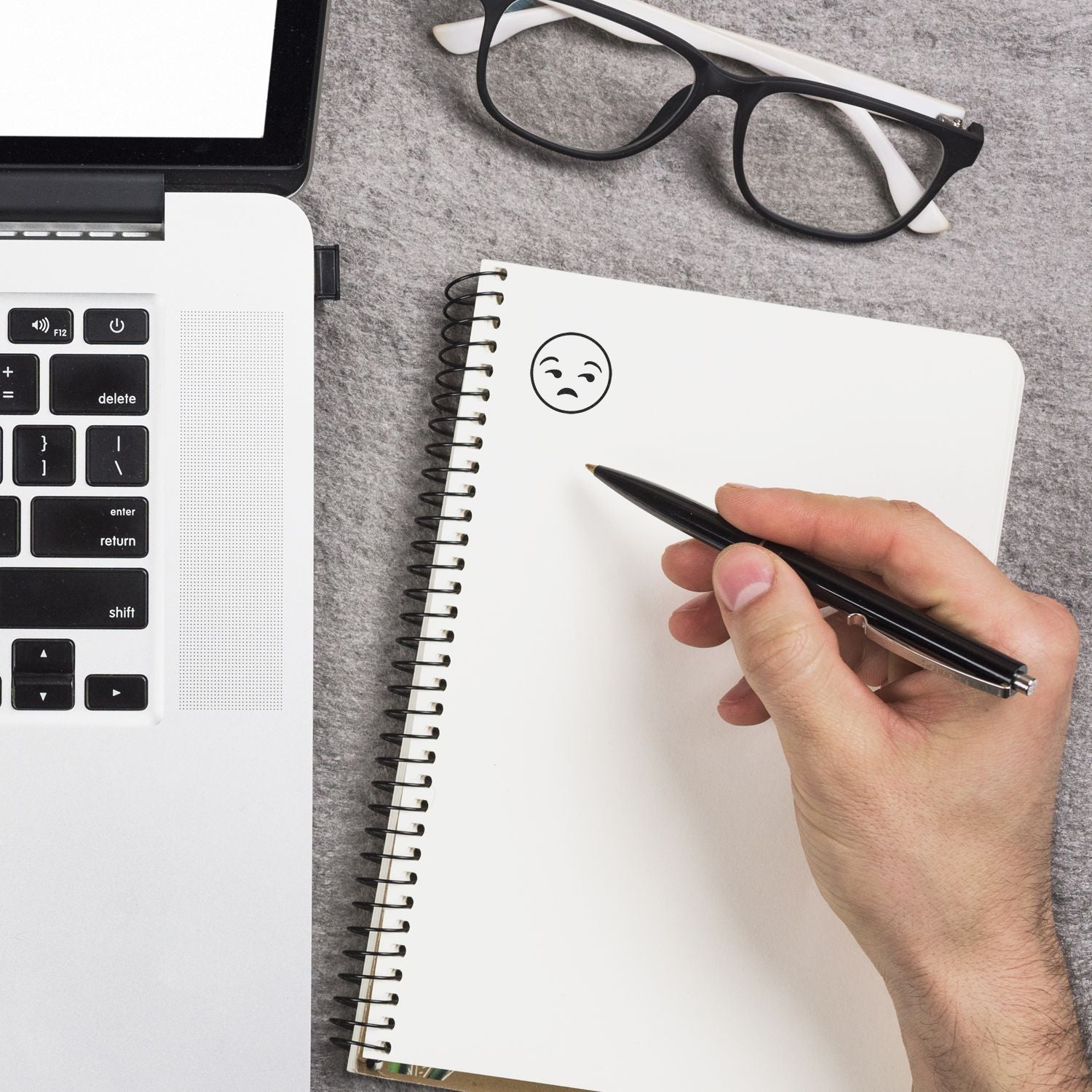 Hand holding pen near notebook with a round calm smiley rubber stamp impression, next to a laptop and glasses.