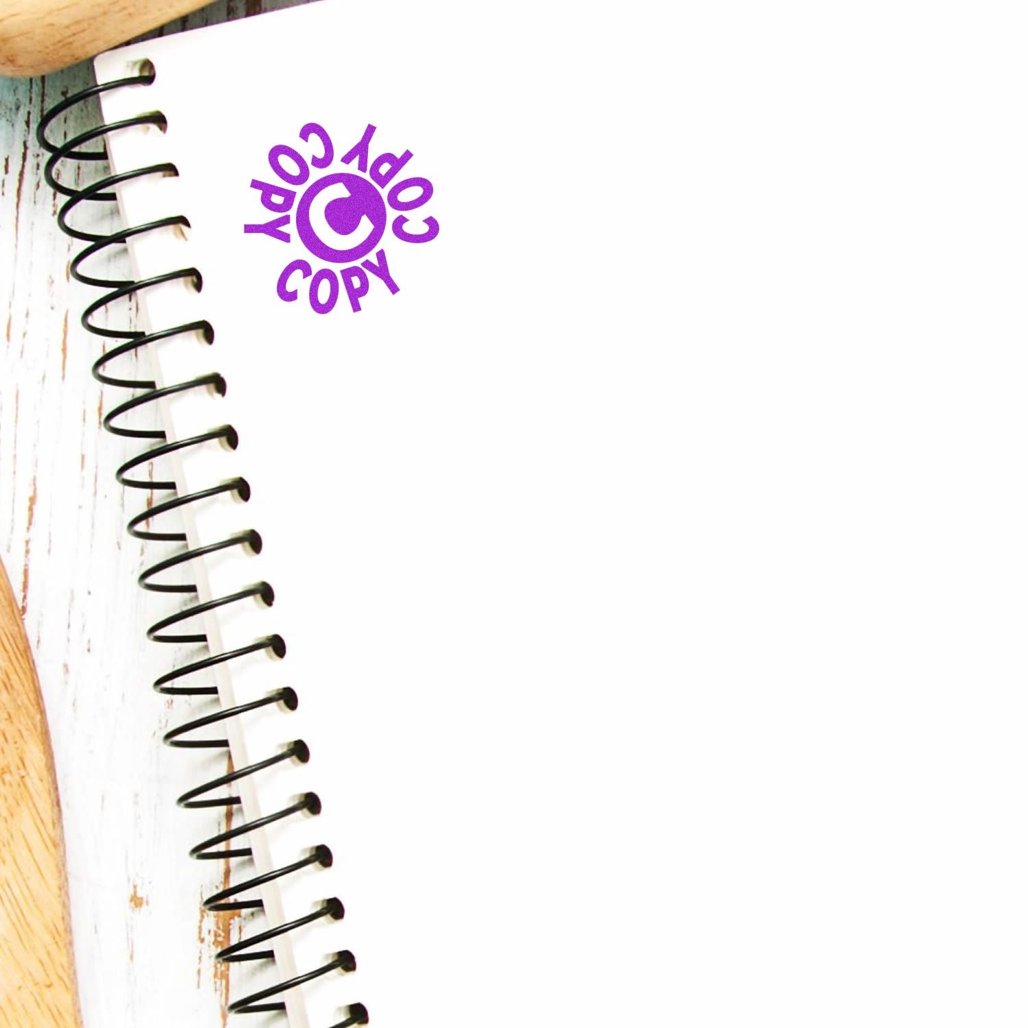 Spiral notebook with a purple Round Copy Copy Copy Rubber Stamp mark on the top left corner, placed on a white wooden surface.