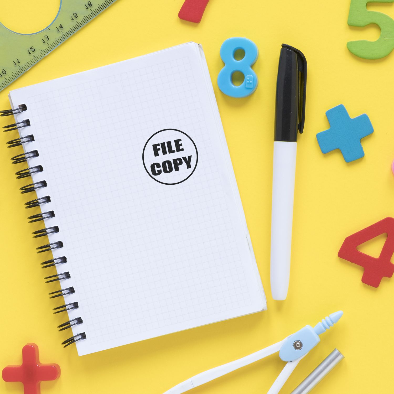 Notebook stamped with Round File Copy Rubber Stamp on a yellow desk with a pen, ruler, and colorful numbers and shapes.