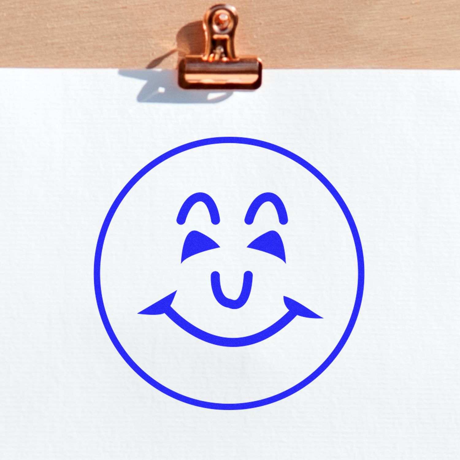 Round Happy Face Rubber Stamp in blue ink on white paper, clipped to a wooden board with a copper clip.