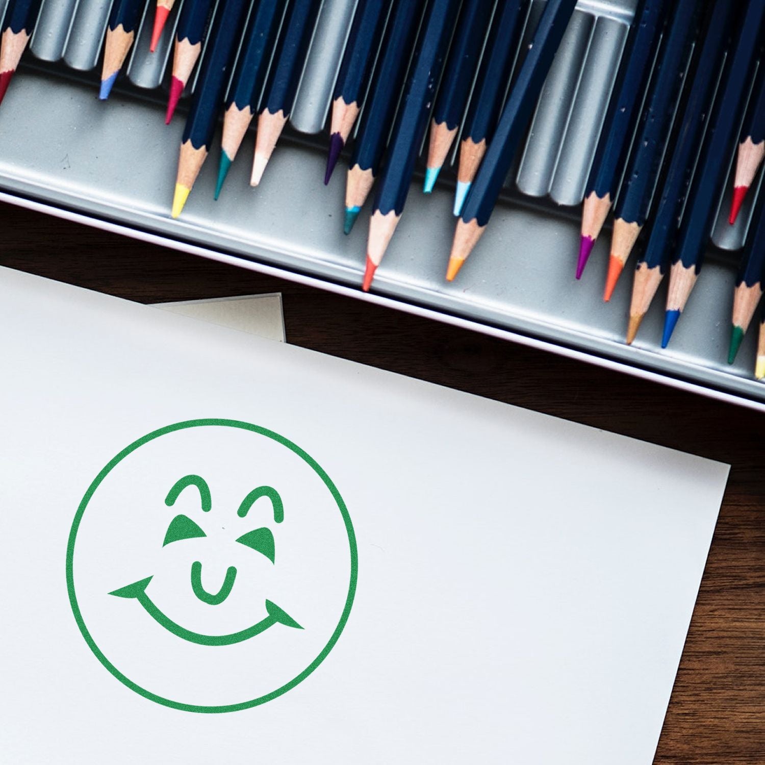 A green Round Happy Face Rubber Stamp impression on white paper, with colored pencils in the background.