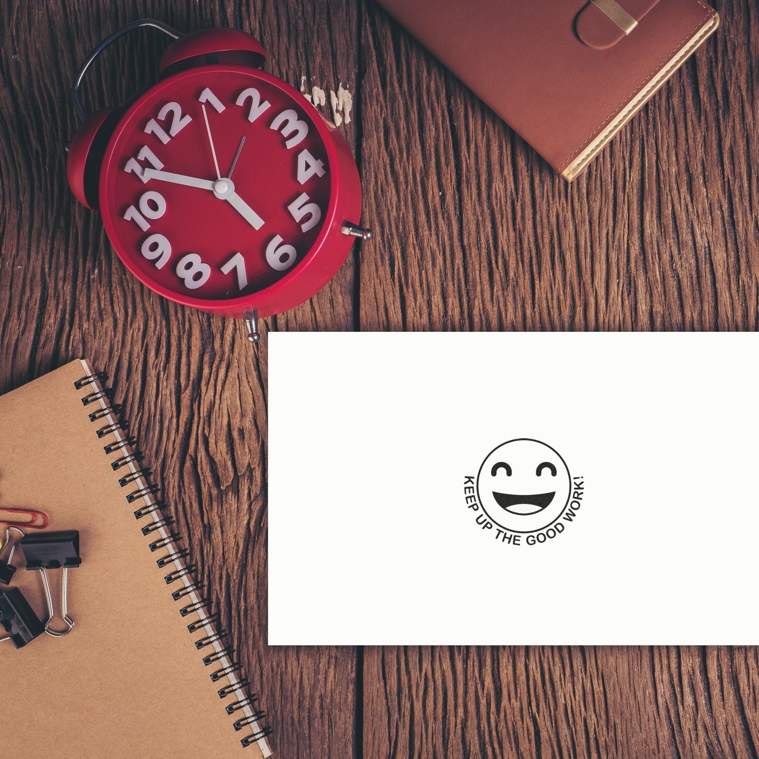 Round Keep up the Good Work Smiley Rubber Stamp on a white paper, placed on a wooden desk with a red clock and notebooks.