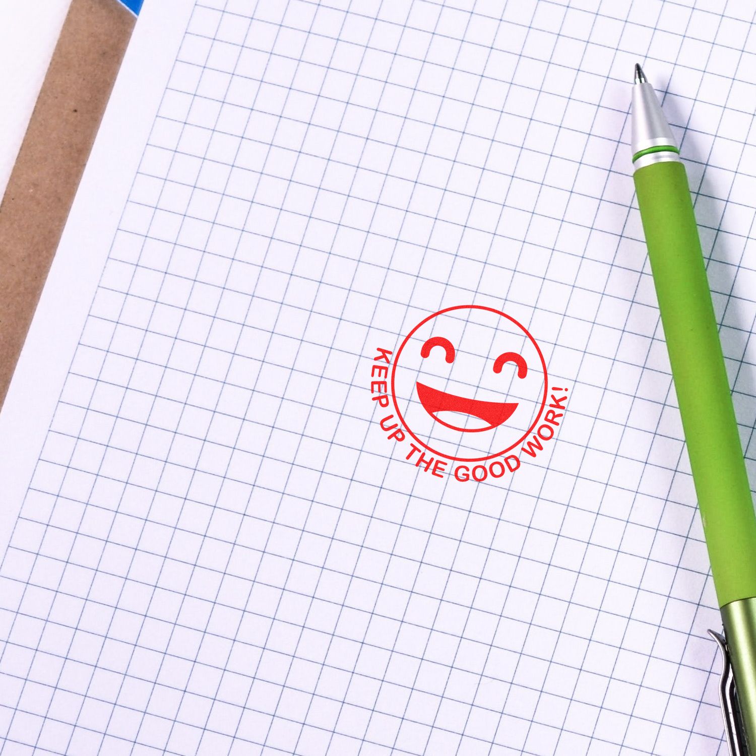 Round Keep up the Good Work Smiley Rubber Stamp in red ink on graph paper, next to a green pen.