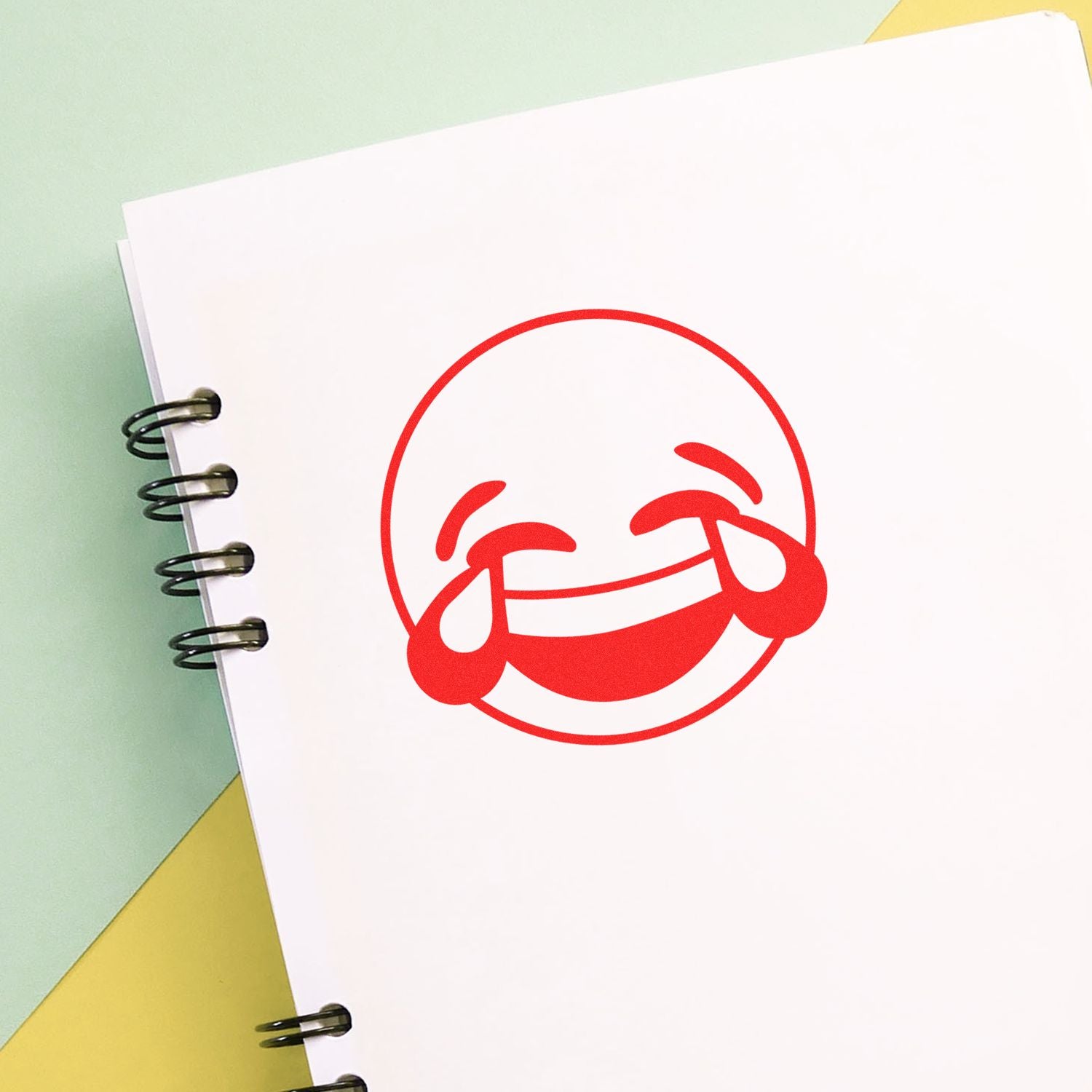 Round Laughing Smiley Rubber Stamp impression on a white spiral notebook page, with a yellow and green background.