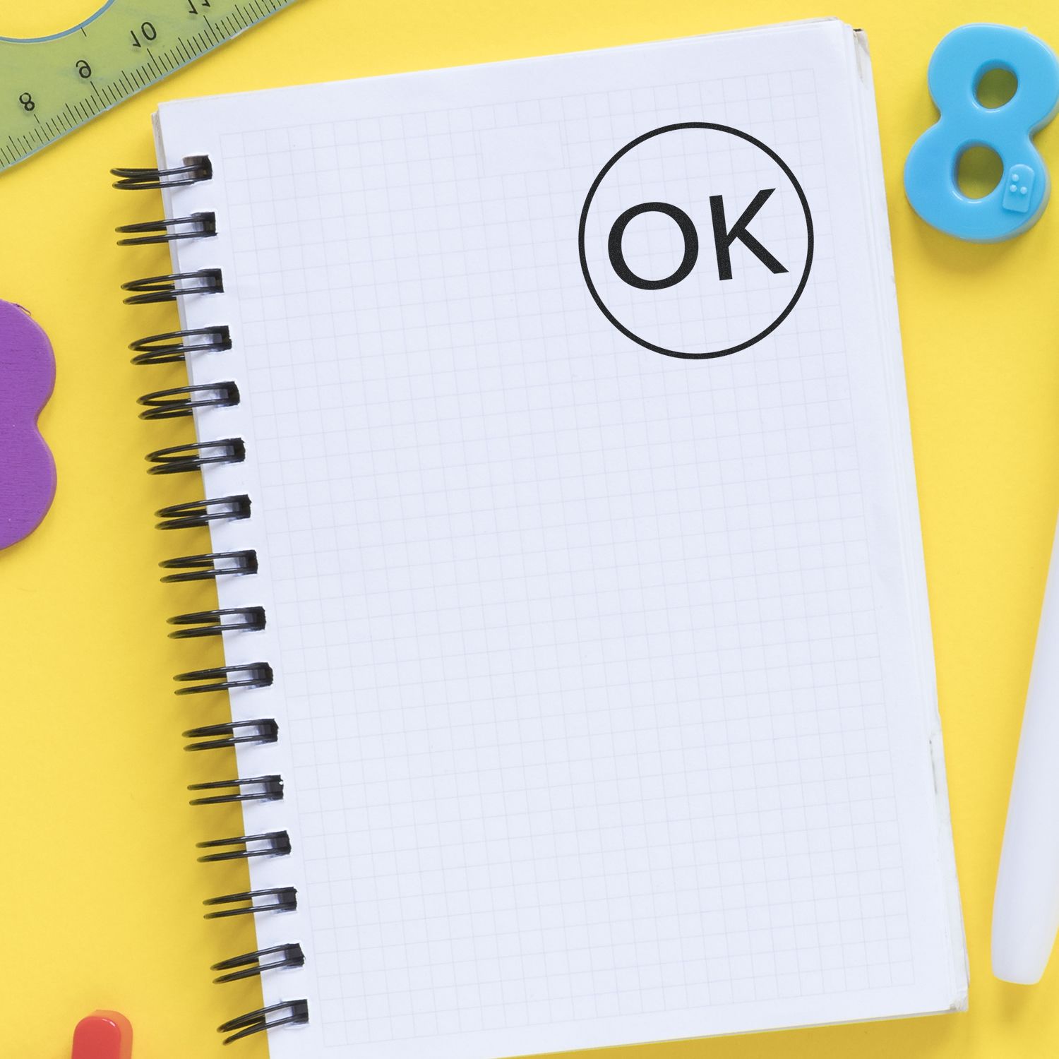 Round OK rubber stamp on a spiral notebook with a grid pattern, surrounded by colorful stationery on a yellow background.