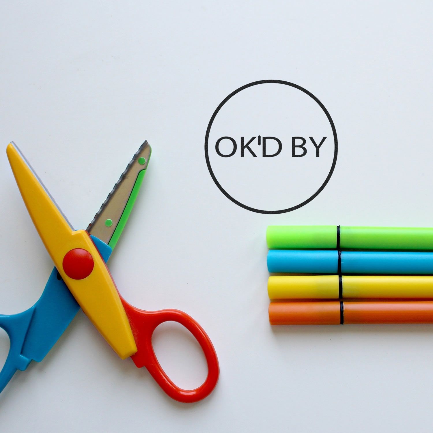 Round OK'd By rubber stamp next to colorful scissors and four stacked markers on a white background.