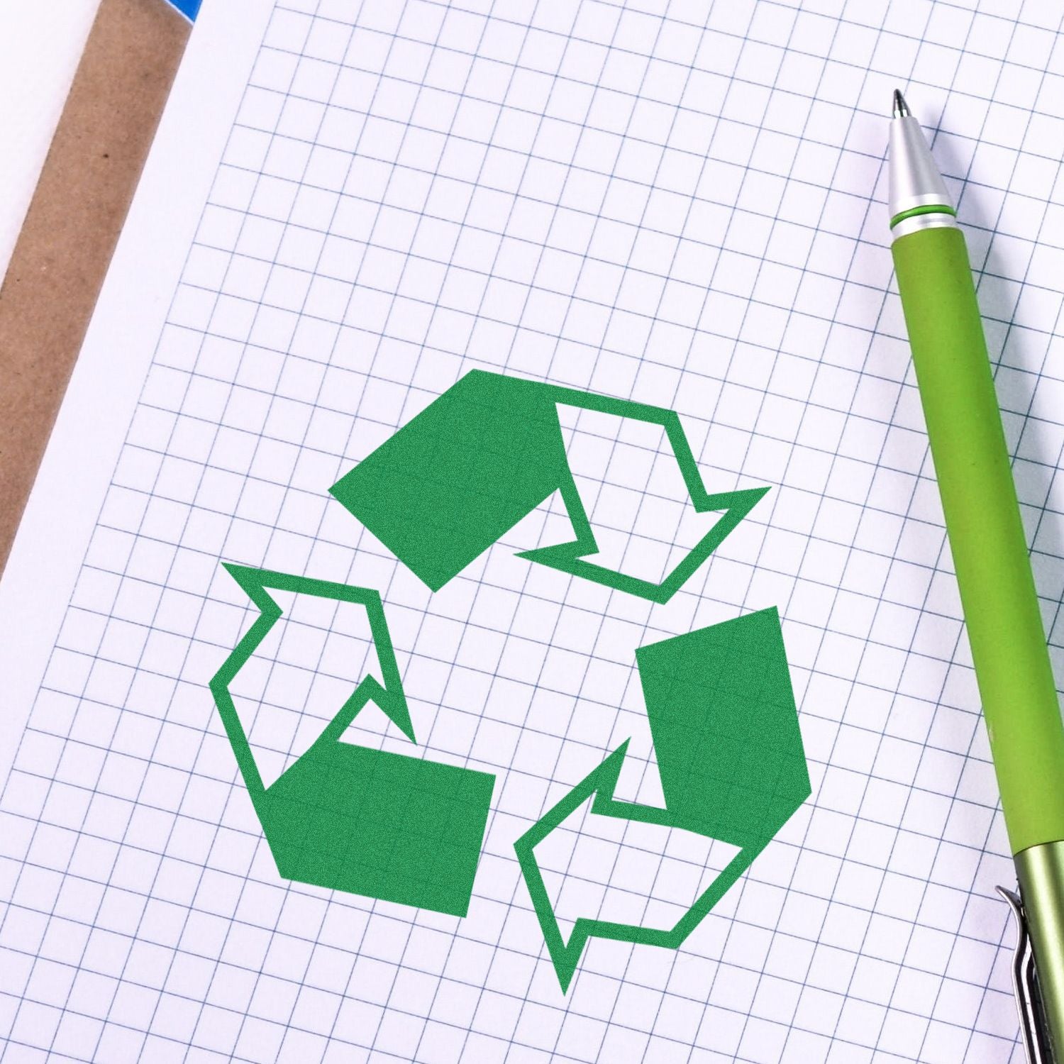 Round Recycle Rubber Stamp in use on graph paper with a green pen beside it. The stamp shows a green recycling symbol.