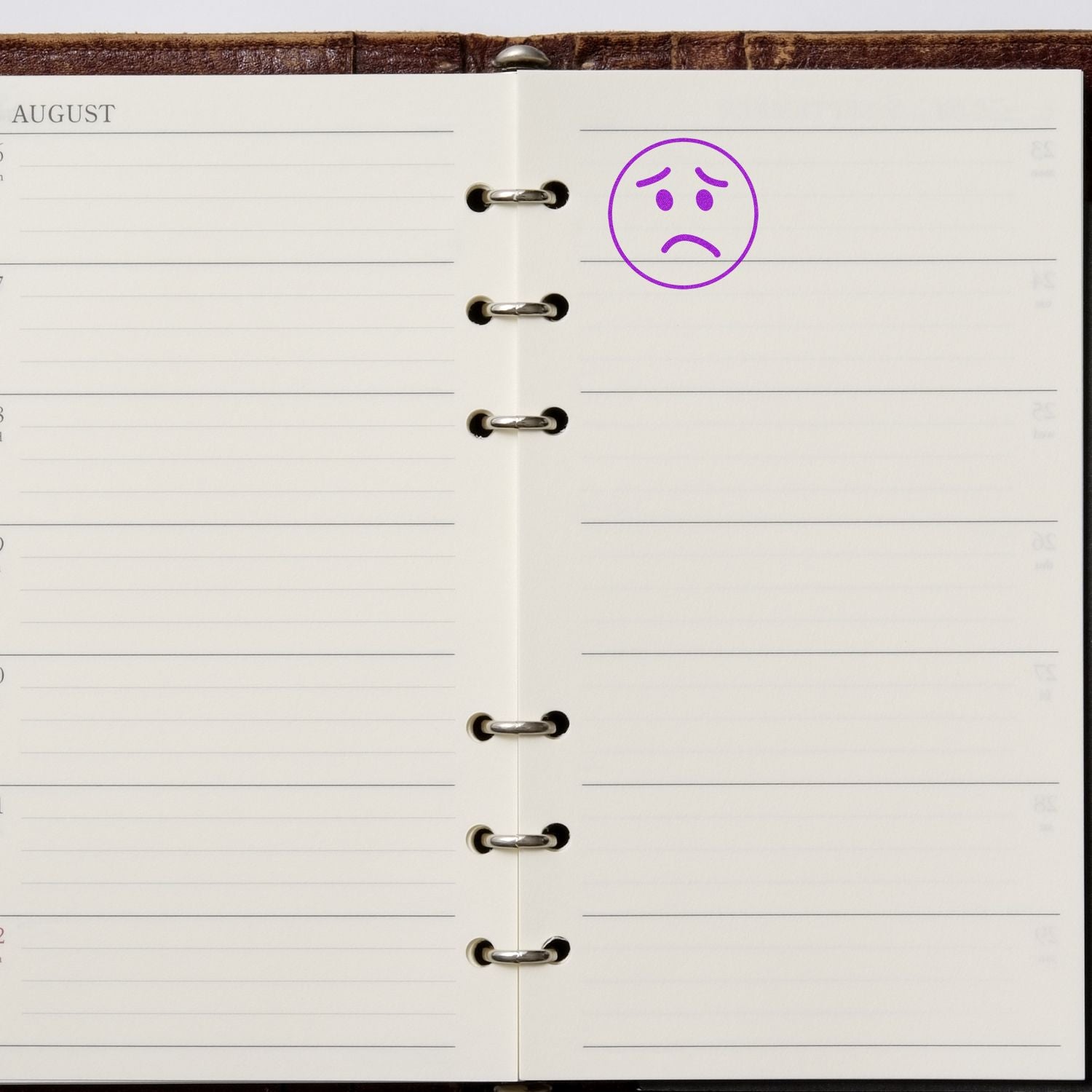 Round Sad Face Rubber Stamp in purple ink on a blank page of an open planner with a brown leather cover.