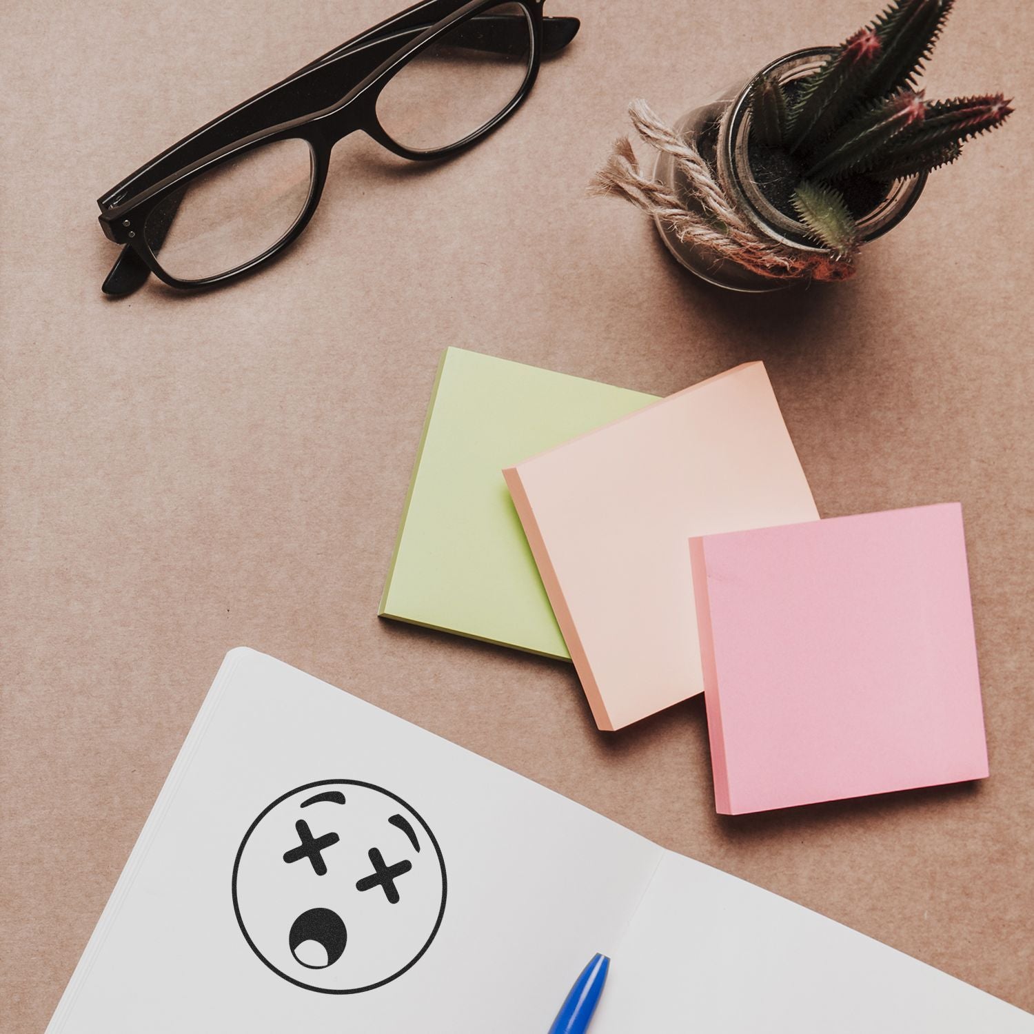 Round Shocked Smiley Rubber Stamp on paper, surrounded by sticky notes, glasses, a pen, and a potted plant on a desk.