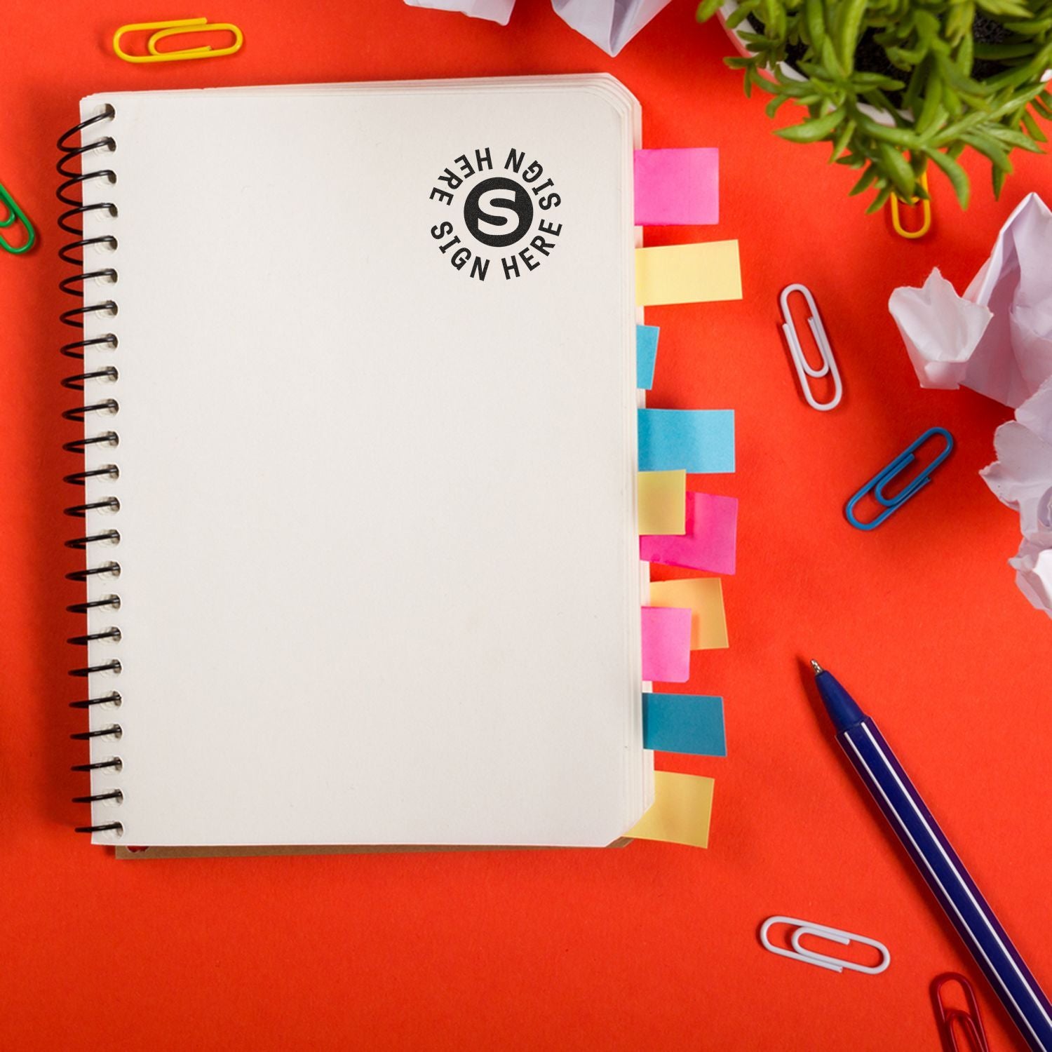 Notebook with colorful tabs and a Round Sign Here Sign Here Rubber Stamp imprint on a red desk with paper clips and a pen.