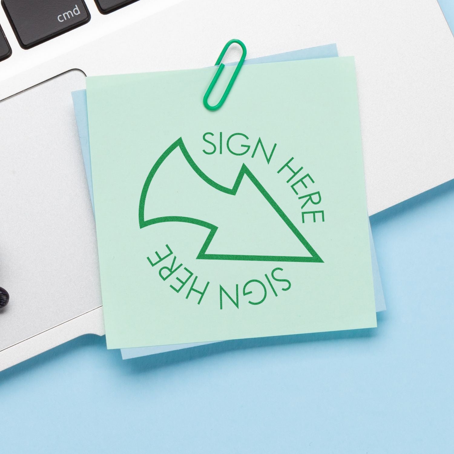 A green Round Sign Here with Arrow rubber stamp on a sticky note, clipped to a laptop keyboard with a paperclip.